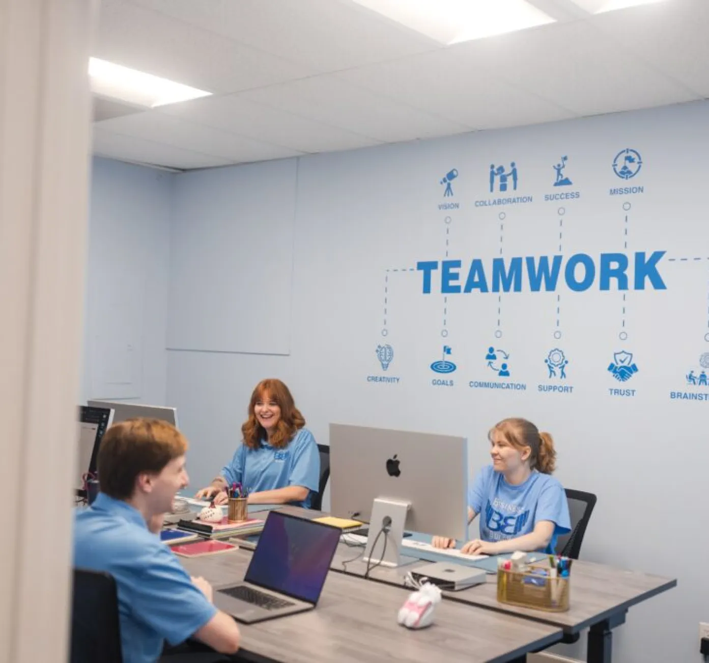 Three people in blue shirts work at desks in an office with a “TEAMWORK” wall graphic and icons behind them.