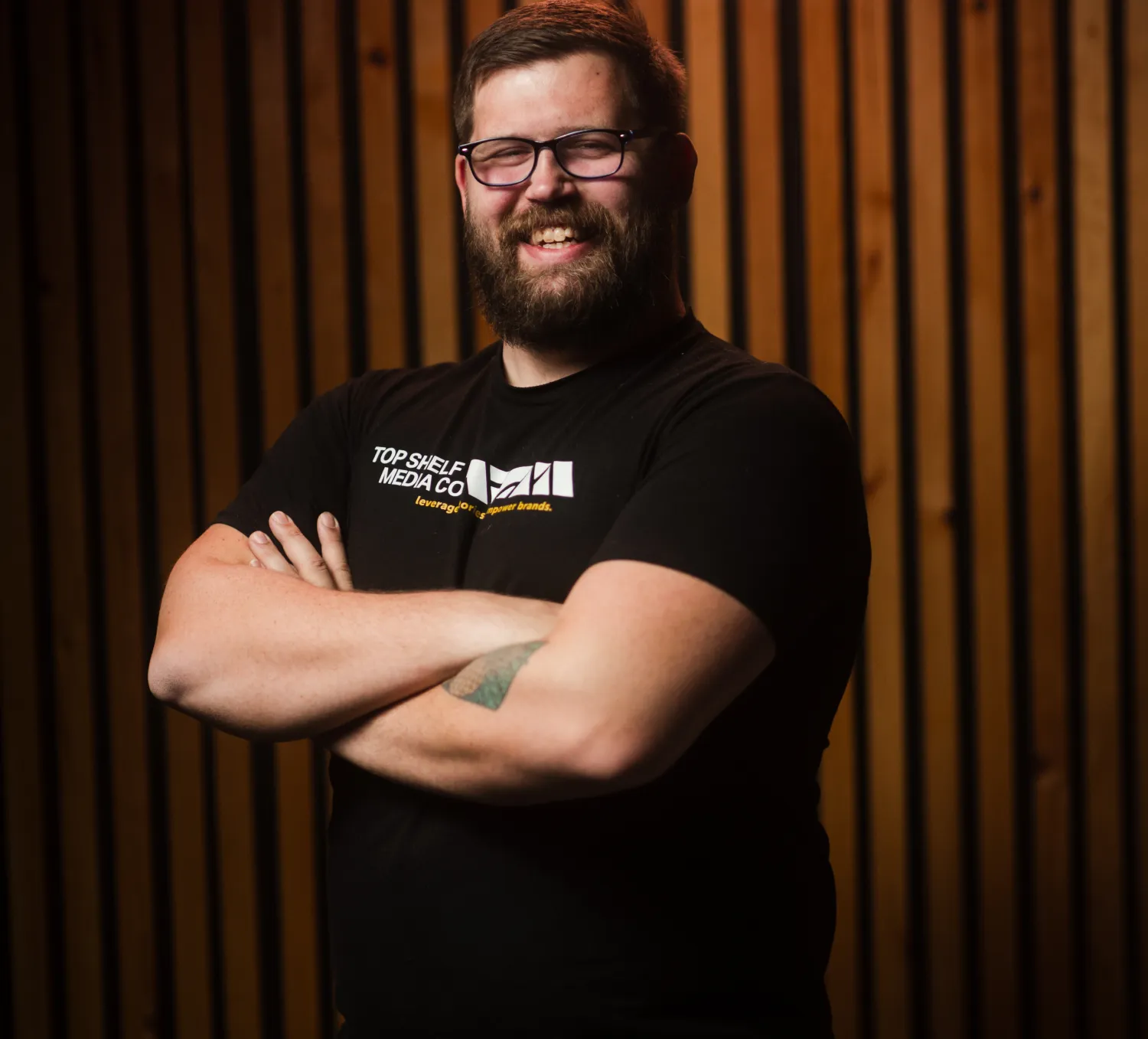 dustin-1 Bearded man with glasses, smiling with arms crossed, stands in front of a wooden slat background.