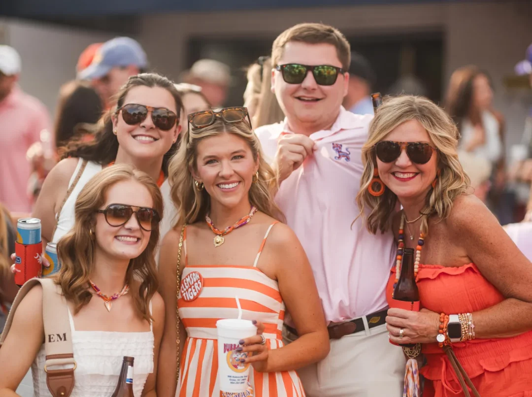 events-13 Five smiling people wearing sunglasses pose outdoors at a festive event, holding drinks and dressed in summer clothes.