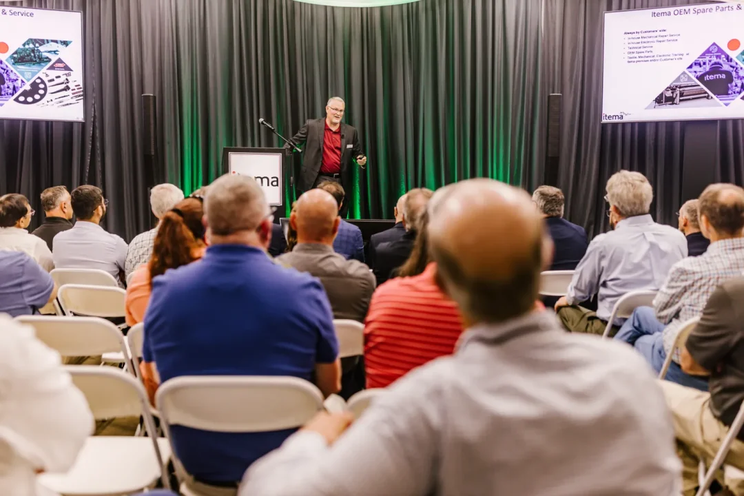 events-19 A speaker presents to a seated audience in a conference room with two display screens and dark curtains.
