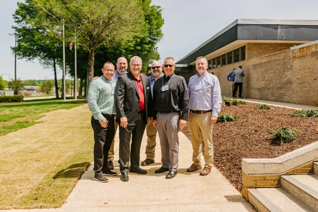 events-20 Six men in business casual attire stand smiling outside near a building on a sunny day.