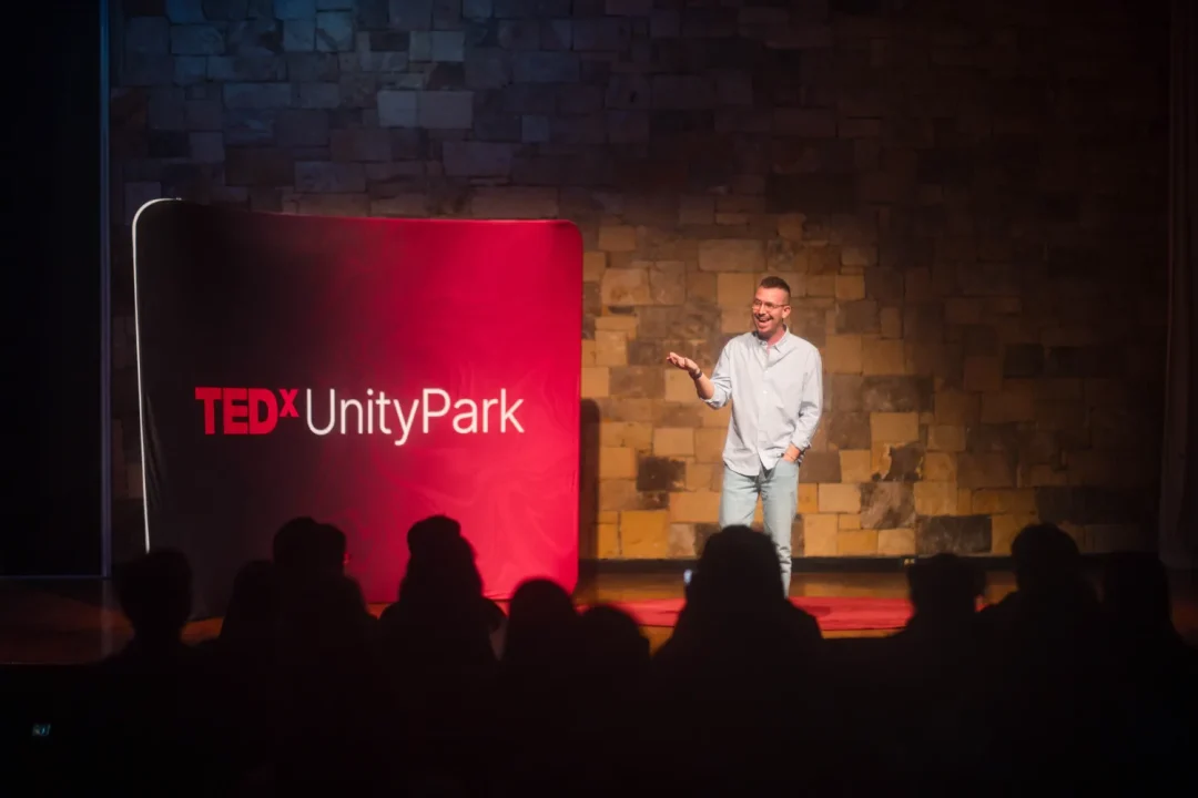 events-28 A speaker stands on stage at TEDxUnityPark, addressing an audience in a dimly lit room.