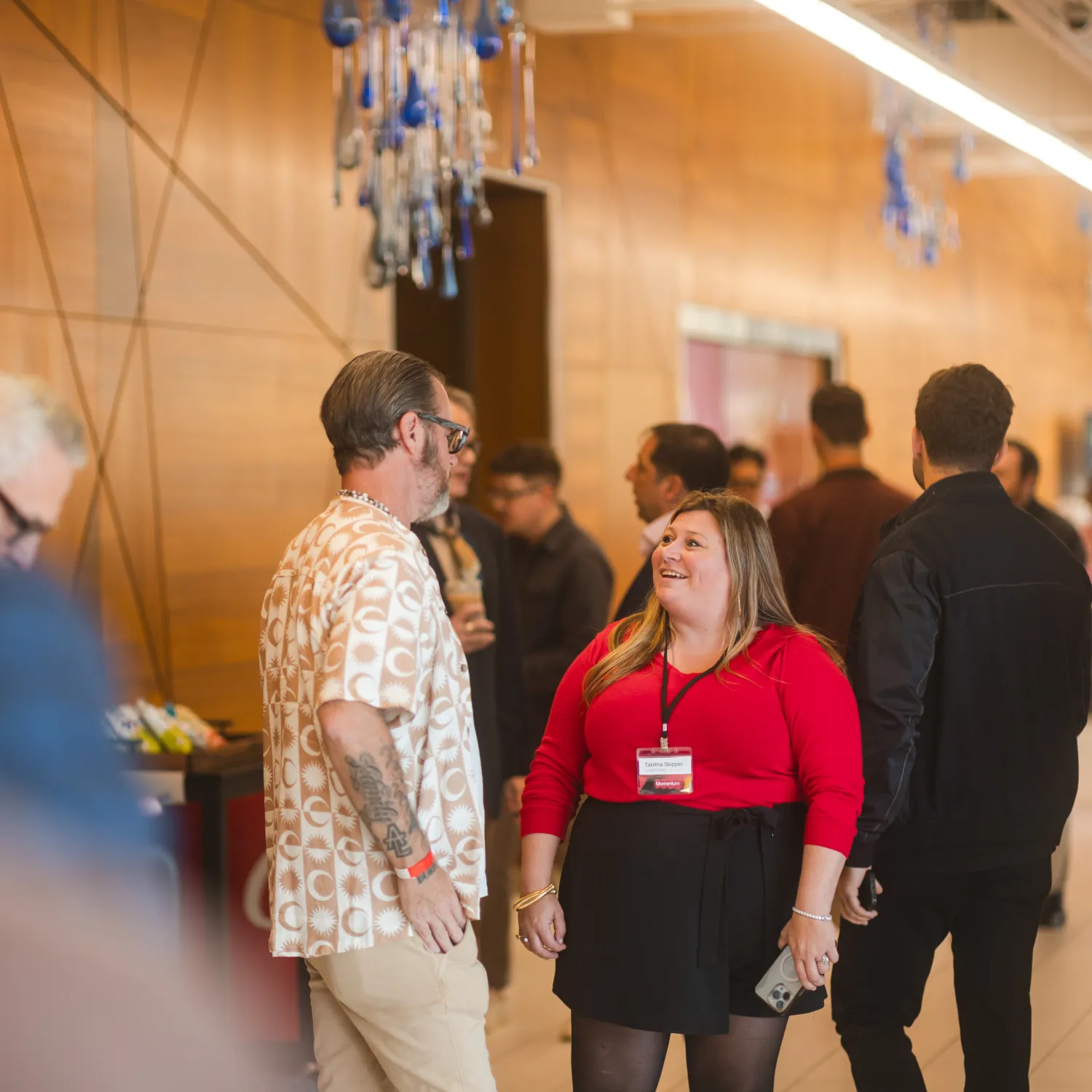 Two people chat at a conference; others mingle in a modern hallway with wood walls and blue decorations.