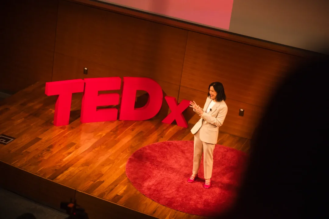 events-5 Speaker in a white suit stands on stage next to large red “TEDx” sign, giving a presentation.