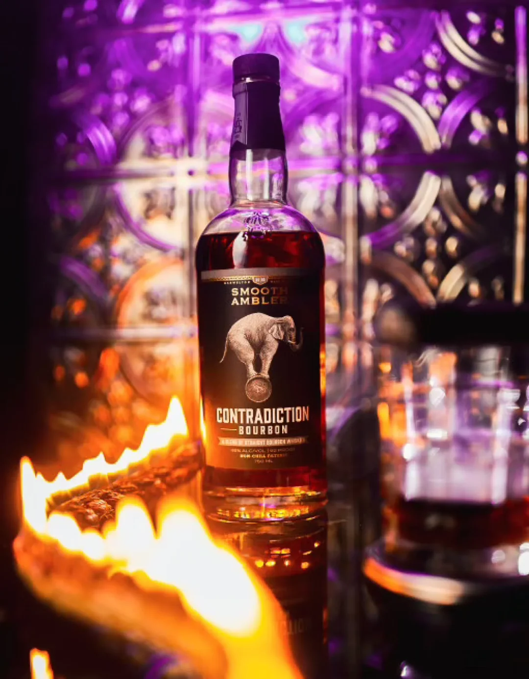 food-10 A bottle of Contradiction Bourbon sits on a bar with purple lighting and fiery orange flames in front.