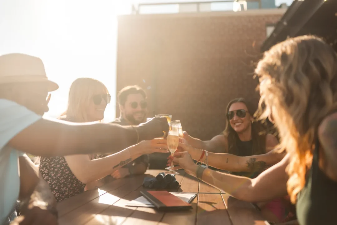 food-5 Friends clink glasses and smile while sitting outdoors at a sunny wooden table.