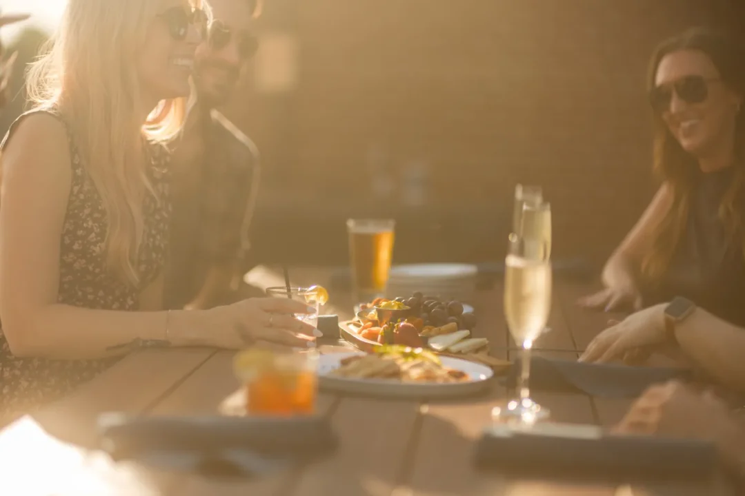 food-7 Three people with drinks and food smile and talk at an outdoor table in warm, golden sunlight.