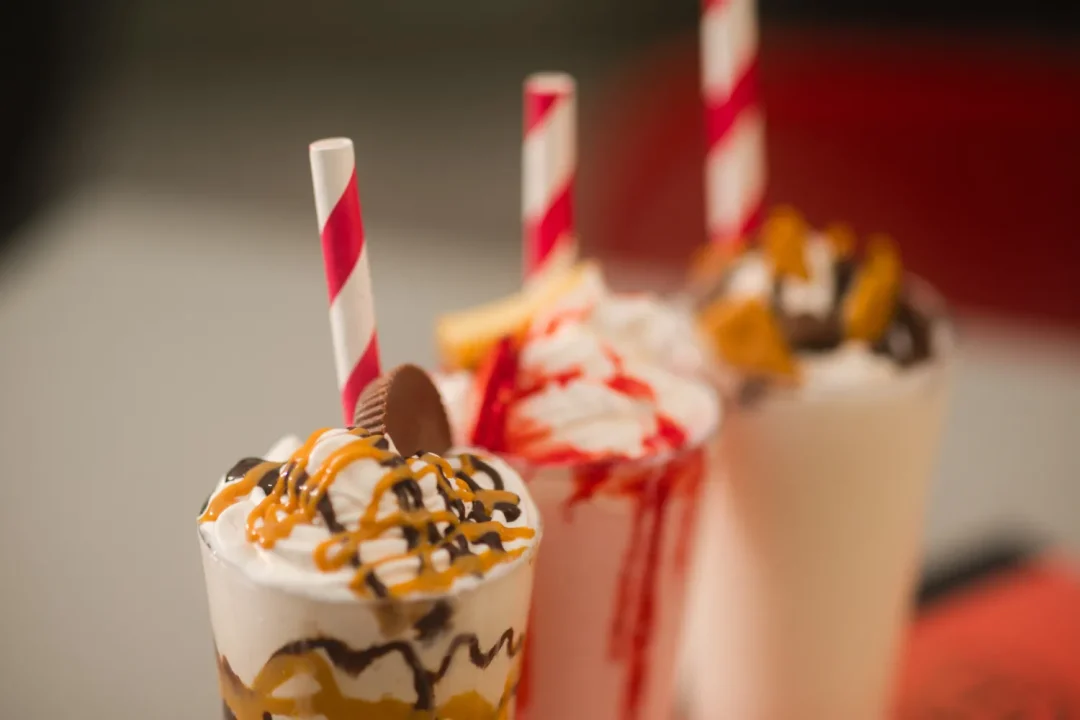 food-8 Three milkshakes with whipped cream, chocolate, caramel drizzle, and red-striped straws, in a row.