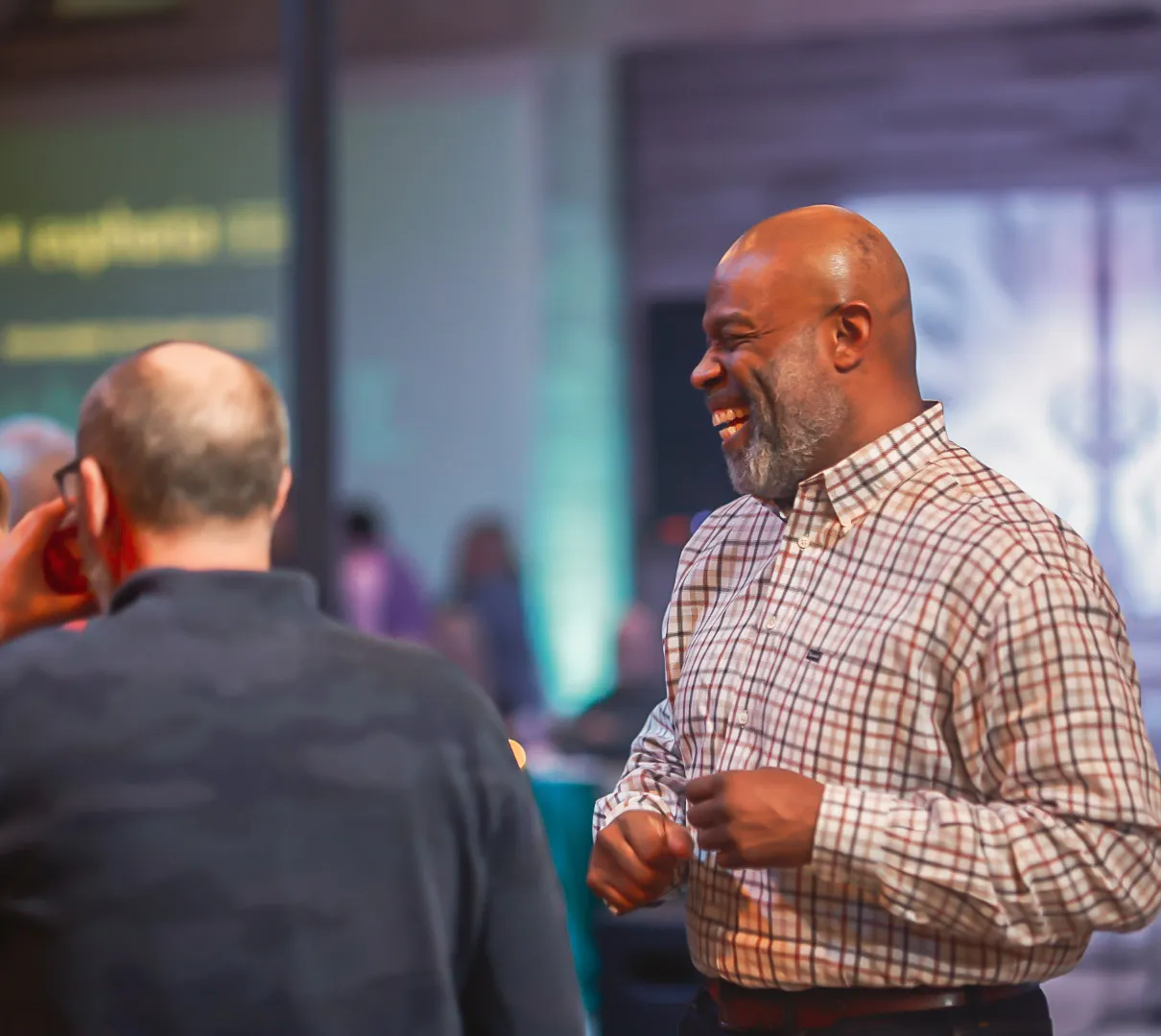 Two men are talking and smiling at an indoor event with colorful lights in the background.