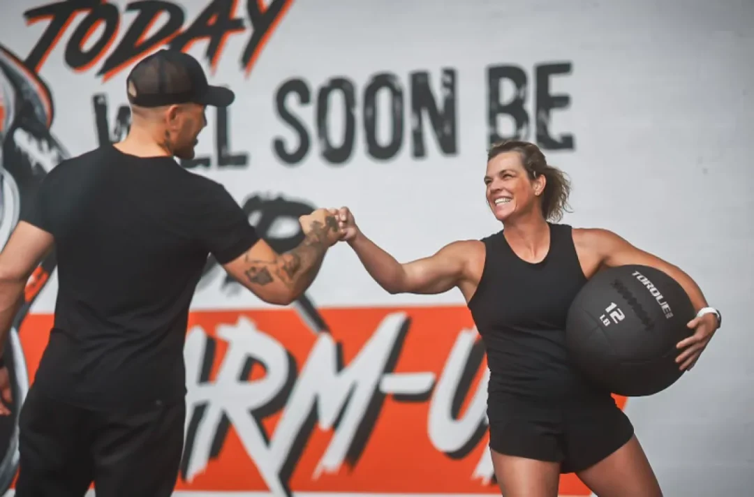 health-wellness-22 Two people fist bump in front of a gym mural; one holds a 12-lb medicine ball and smiles.