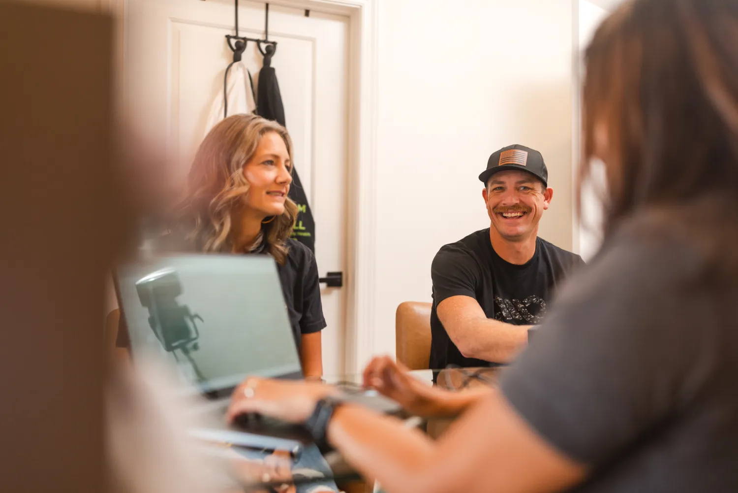 Three people sit around a table, talking and smiling, with a laptop open in the foreground.