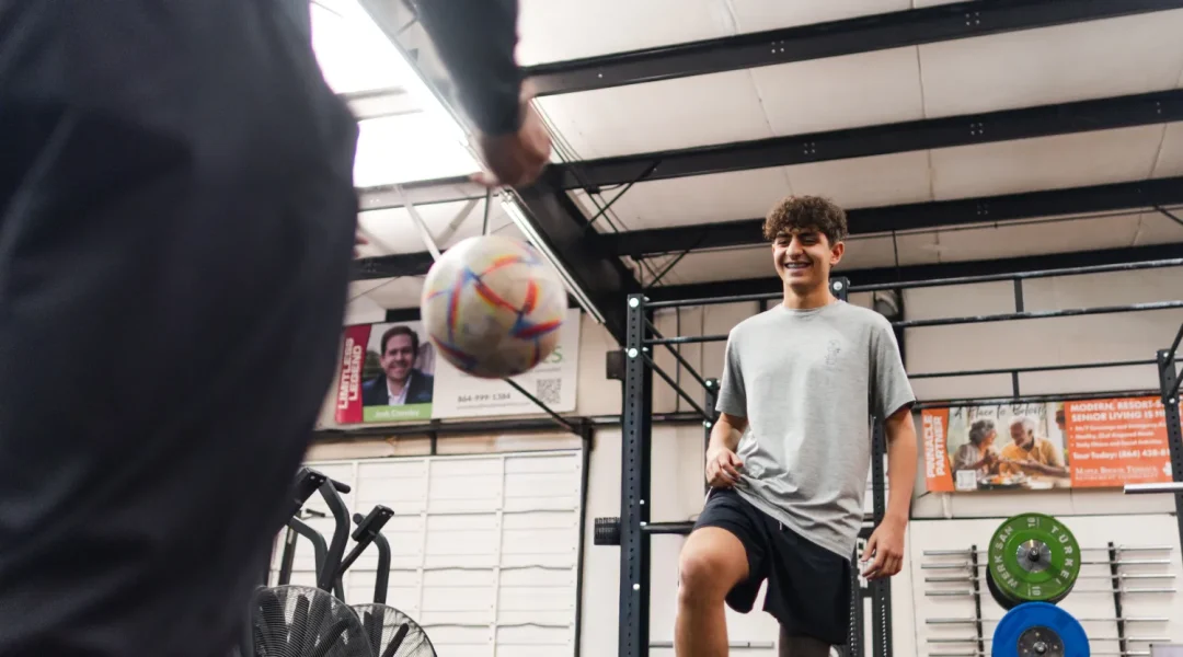 health-wellness-30 Teenage boy smiling while practicing soccer skills indoors with another person in a gym setting.