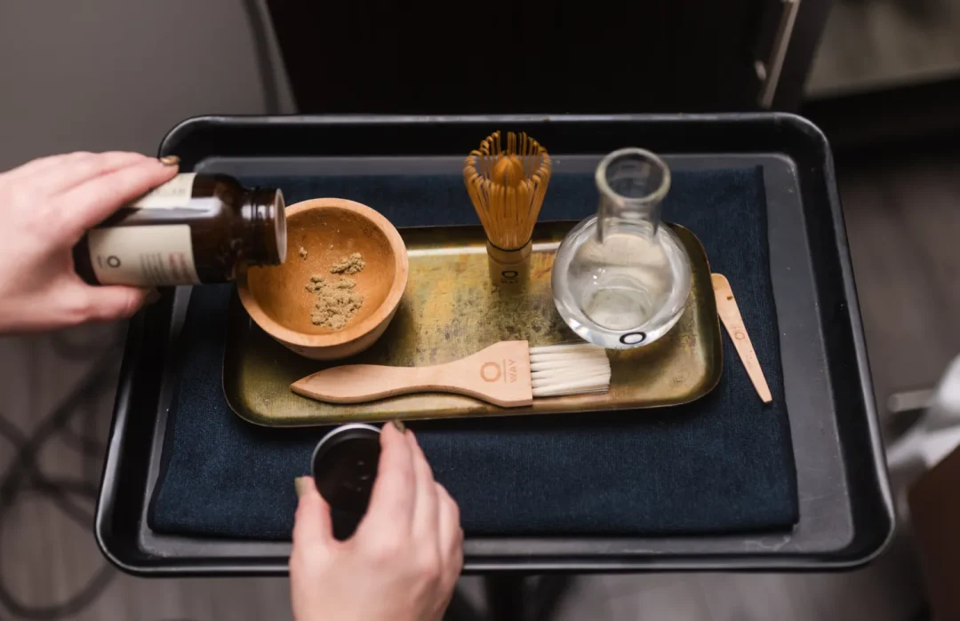 health-wellness-7 Hands preparing a bowl of matcha with tools: whisk, brush, spoon, and glass carafe on a tray.