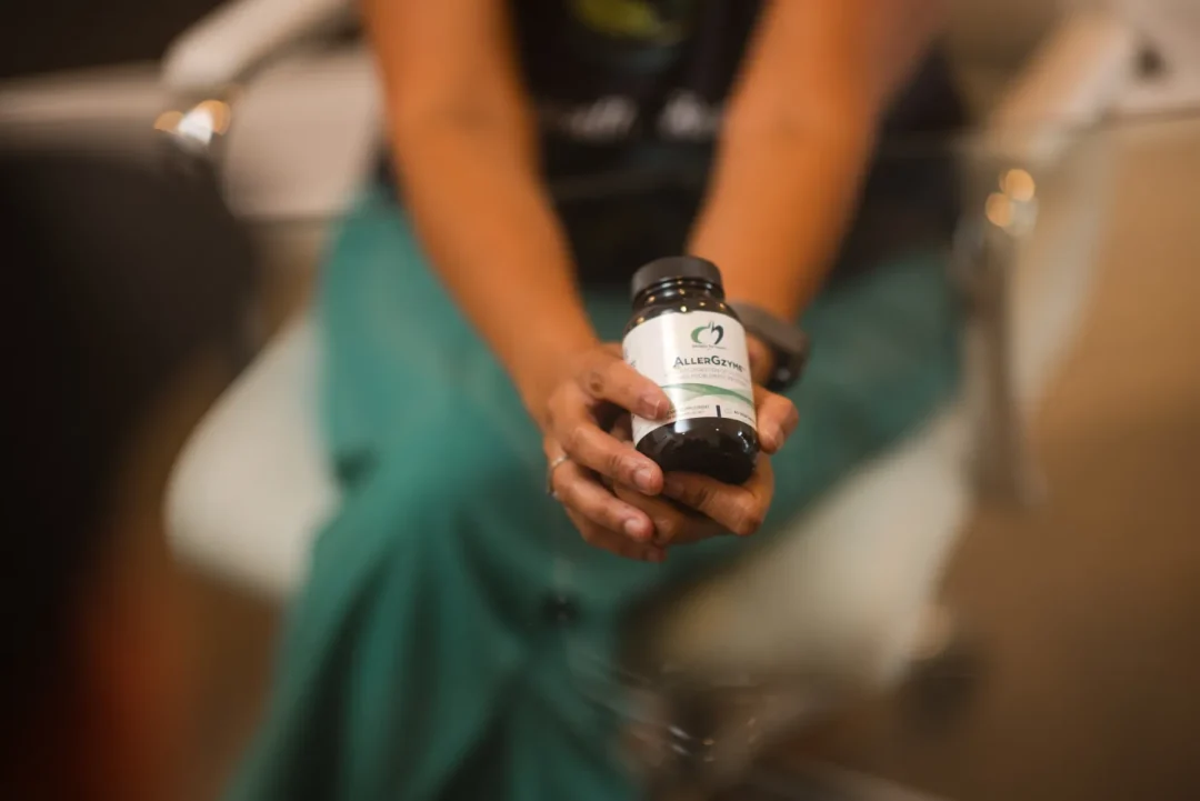 health-wellness-8 Person holding a bottle of supplements labeled “Allugen” while sitting at a glass table.
