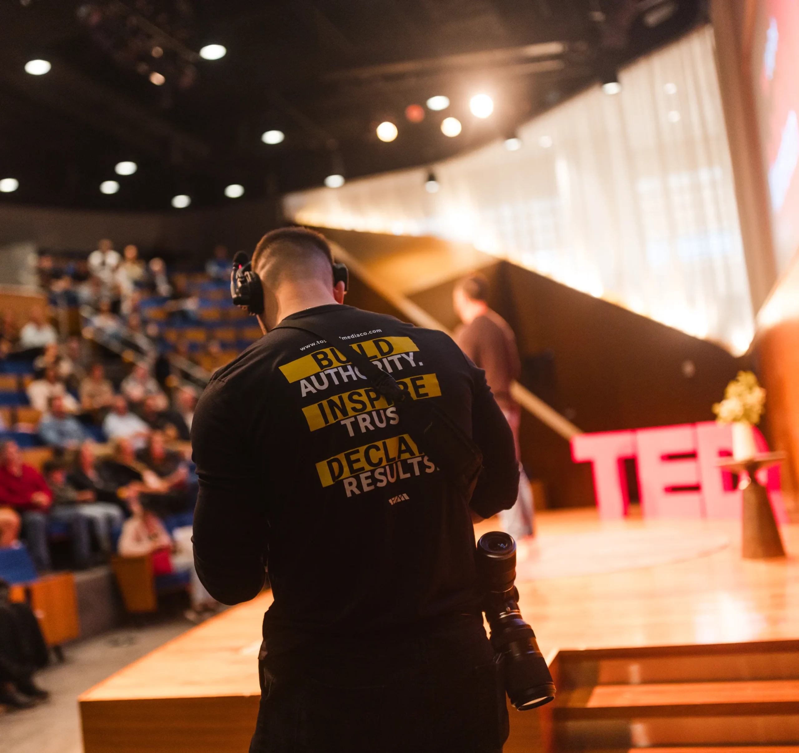 home-1 A photographer with a camera stands near a TED stage as people sit in an auditorium.