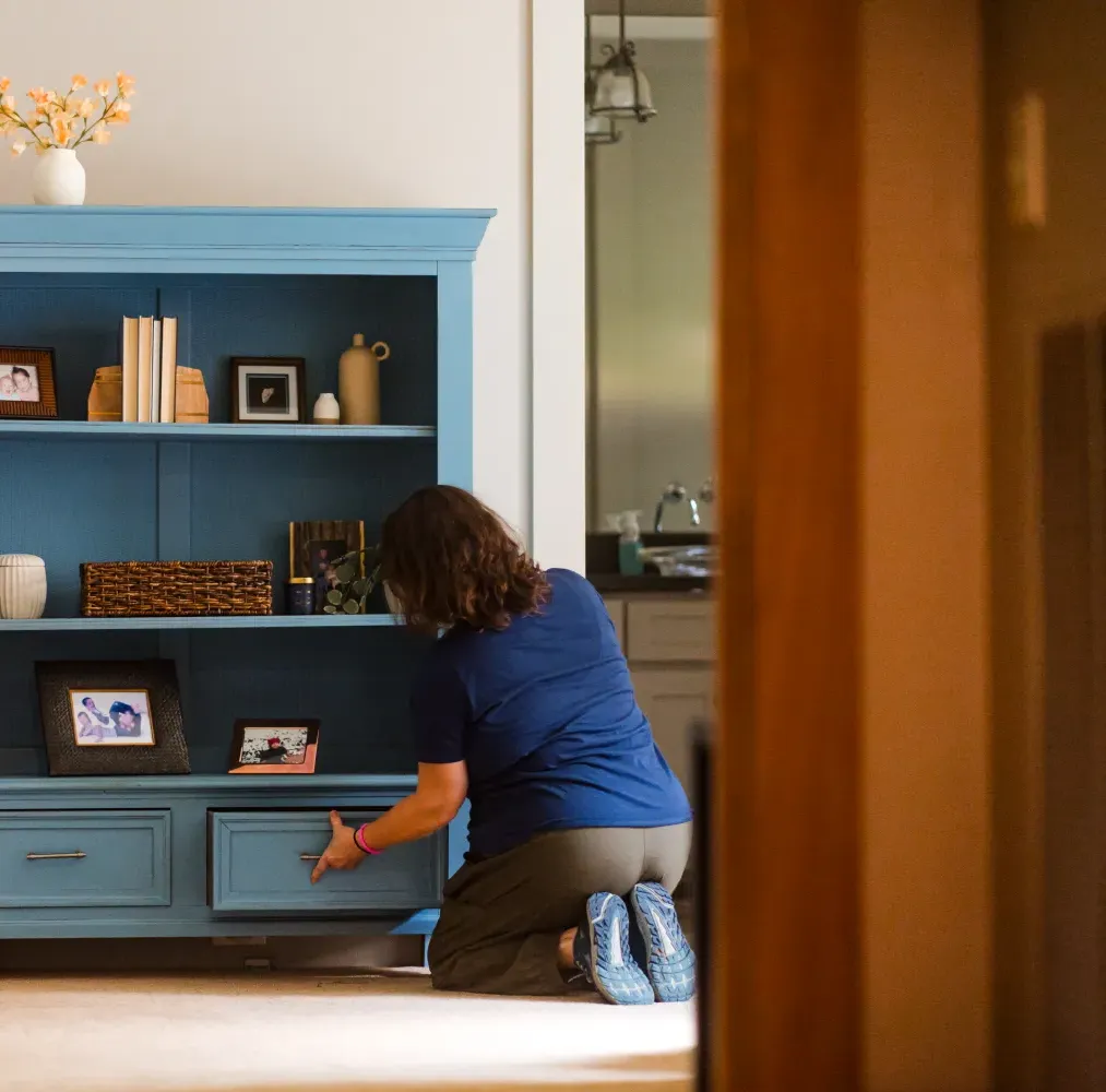 A person kneels on the floor, organizing items in a blue cabinet in a warmly lit room.
