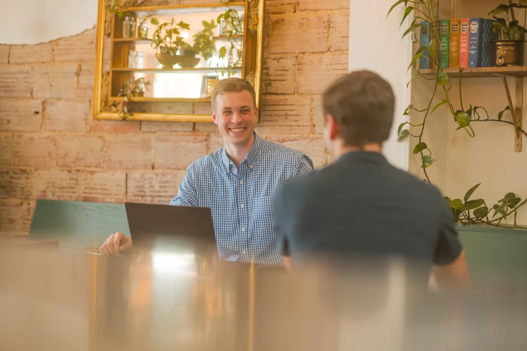 professional-1 Two people sit at a table in a cozy cafe, one smiling with a laptop, plants on shelves behind them.