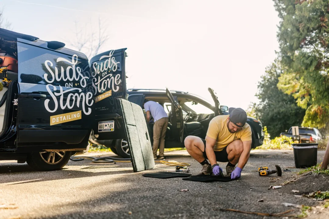 professional-14 Man in gloves cleans car mats near a van labeled “Suds on Stone Detailing” on a sunny day.