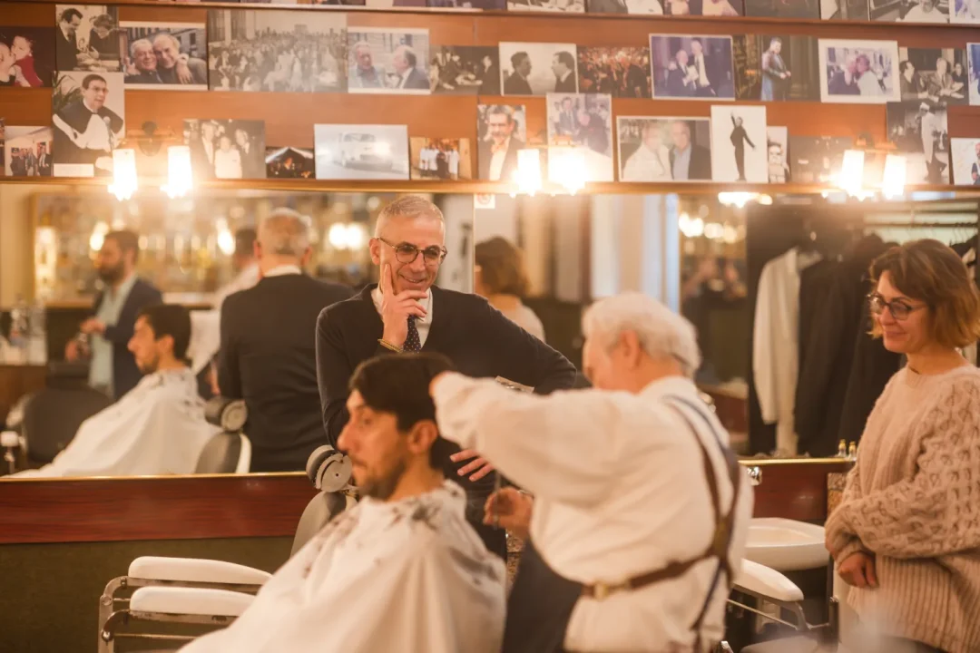 professional-2 A barber cuts a man’s hair while two others watch in a warmly lit barbershop with photos on the wall.