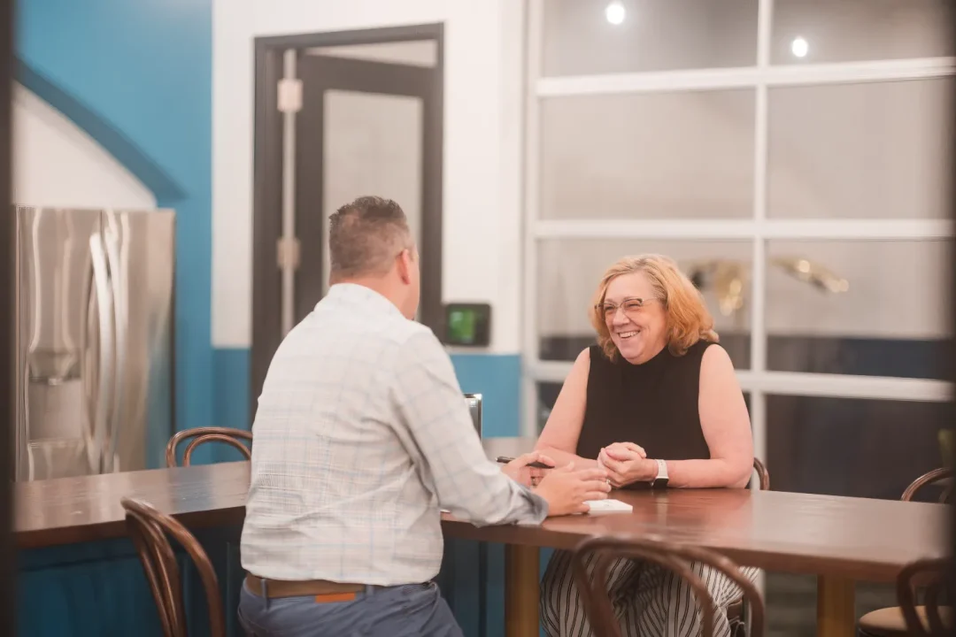 professional-5 Two people sit at a table in conversation, smiling in a modern indoor space.