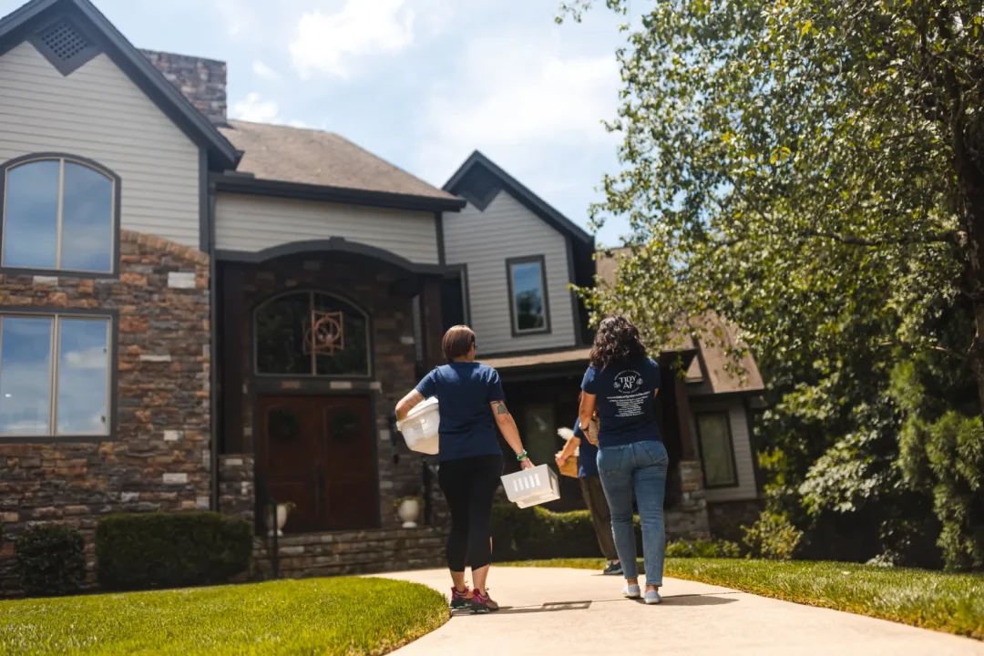 professional-6 Two people carrying cleaning supplies walk toward the front door of a large stone and wood house.