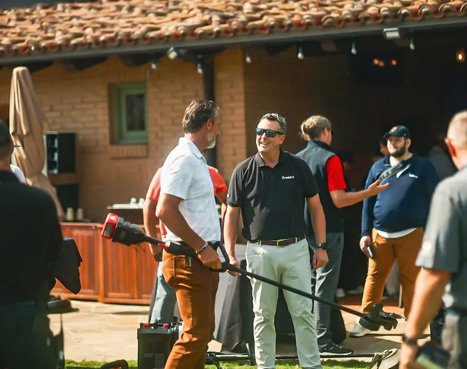 Two men talk outside at an event, one holding equipment, others mingle in the sunny courtyard.