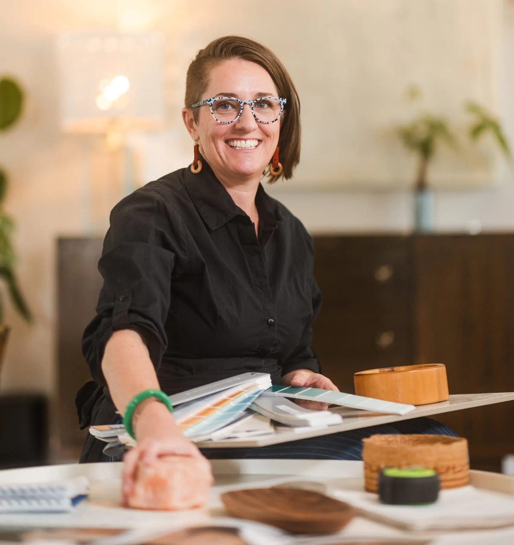 Smiling person with glasses sits at a table with books, samples, and wooden objects in a well-lit room.