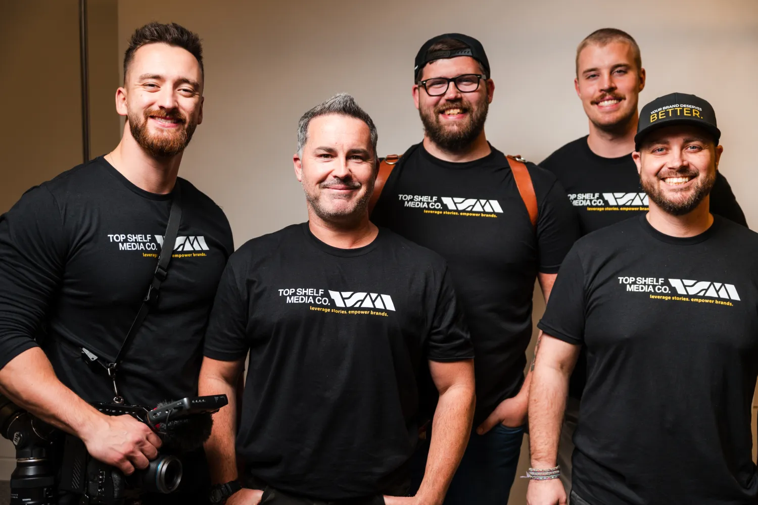 team-1 Five men in matching black shirts, one holding a camera, smiling and posing together indoors.
