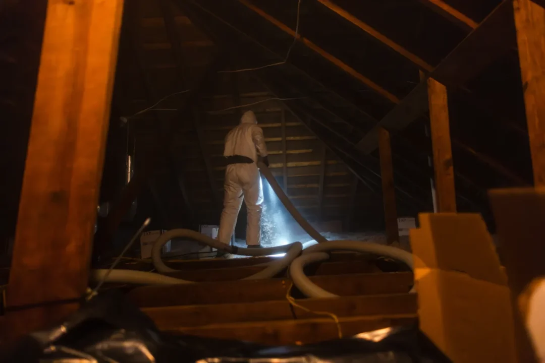trades-10 Person in protective suit sprays insulation in a dimly lit attic with exposed beams and hoses.