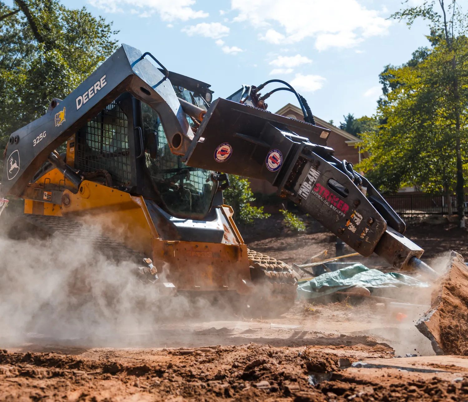 trades-14 A John Deere skid steer uses a hydraulic attachment to break concrete, kicking up dust at a work site.