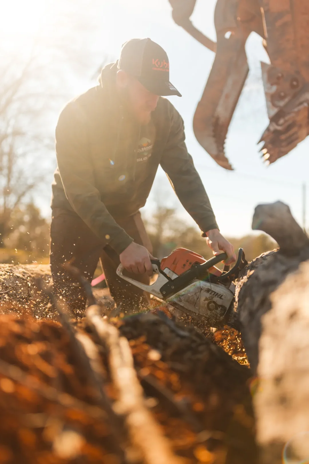 trades-18 Person using a chainsaw to cut a log outdoors, with wood chips flying in bright sunlight.