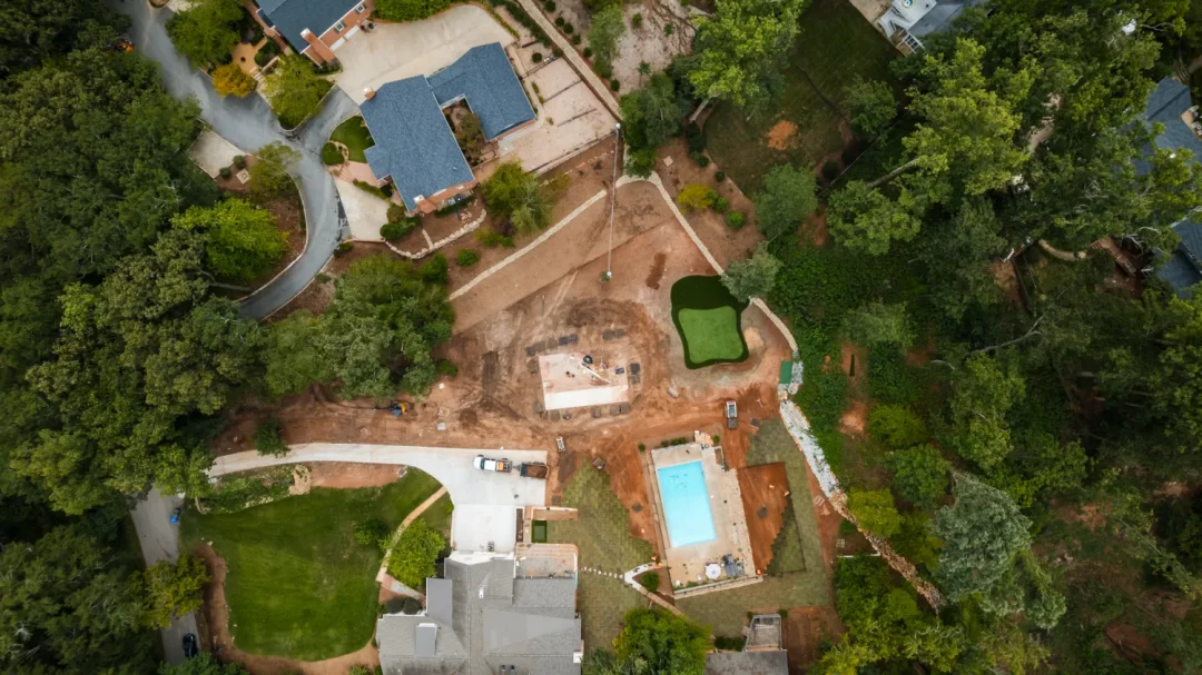 trades-30 Aerial view of a residential area with houses, a swimming pool, dirt lots, and surrounding trees.