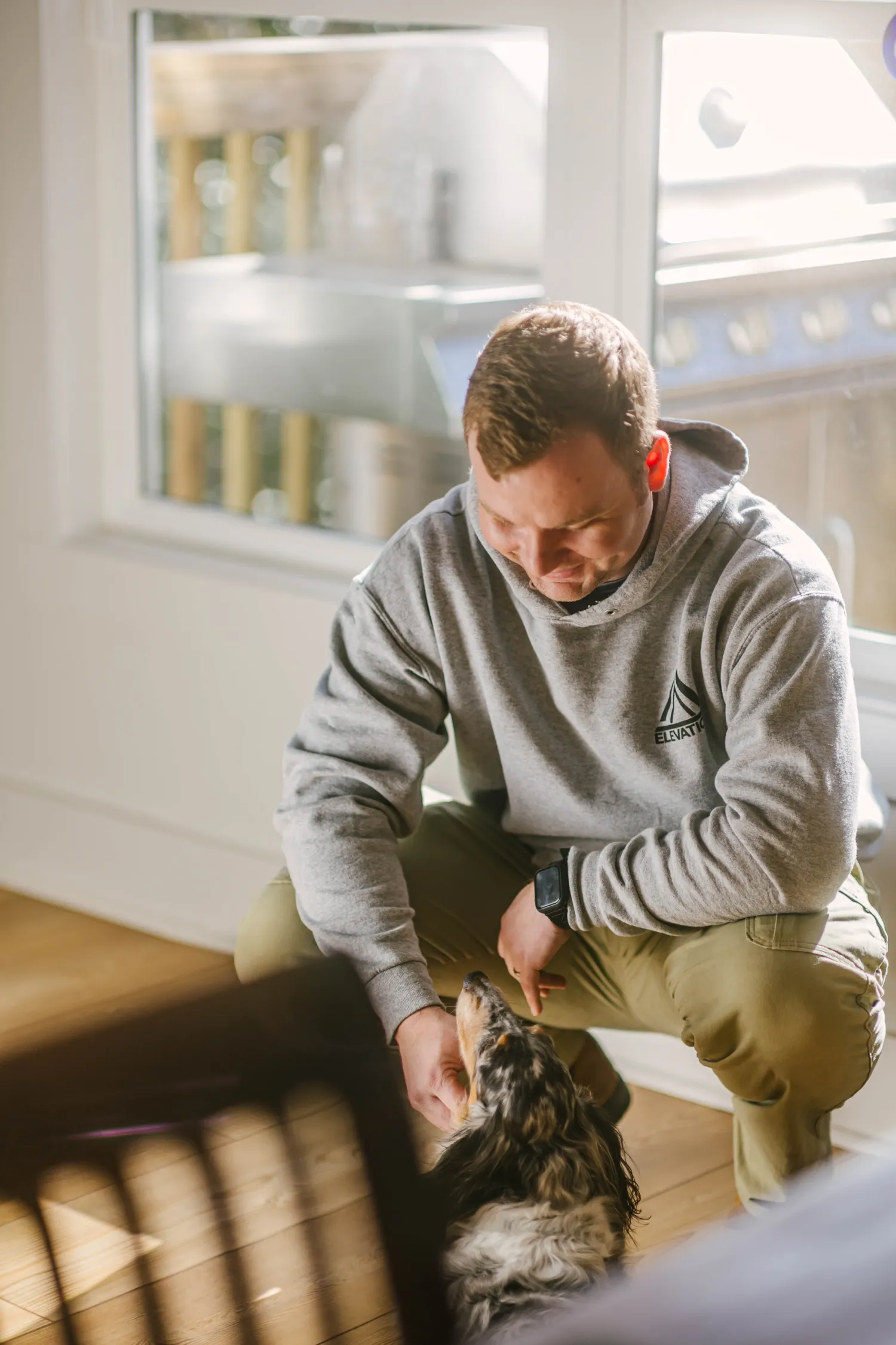 trades-36 Man in a gray hoodie kneels indoors, smiling and petting a dog near a sunlit window.