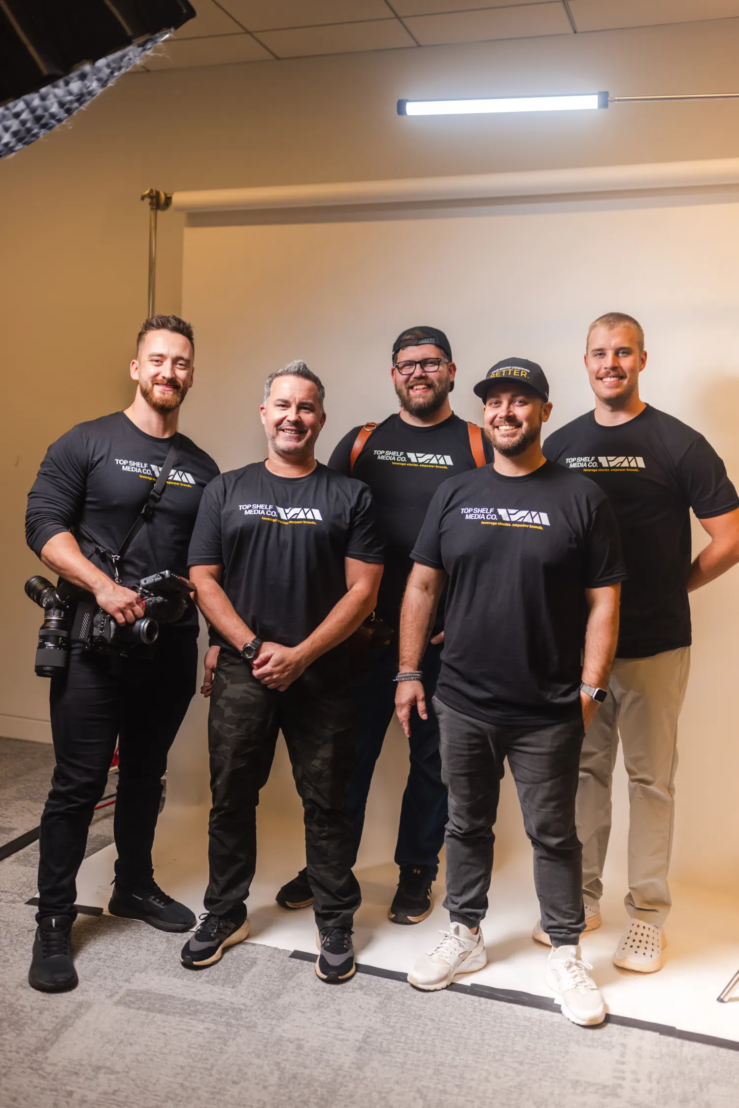 why-choose-1 Five men in black shirts posing and smiling together in a photography studio with a white backdrop.