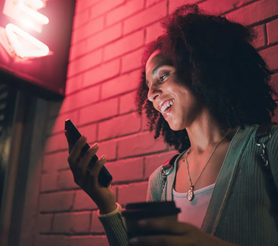 who-1 Smiling woman holding coffee and looking at her phone under pink neon lights near a brick wall at night.