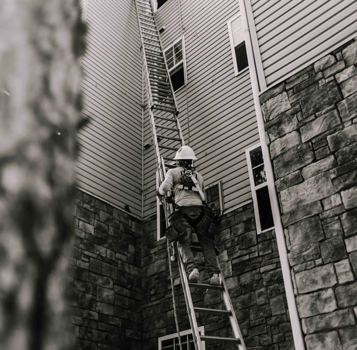 A construction worker climbs a ladder beside a tall building with stone and siding walls.