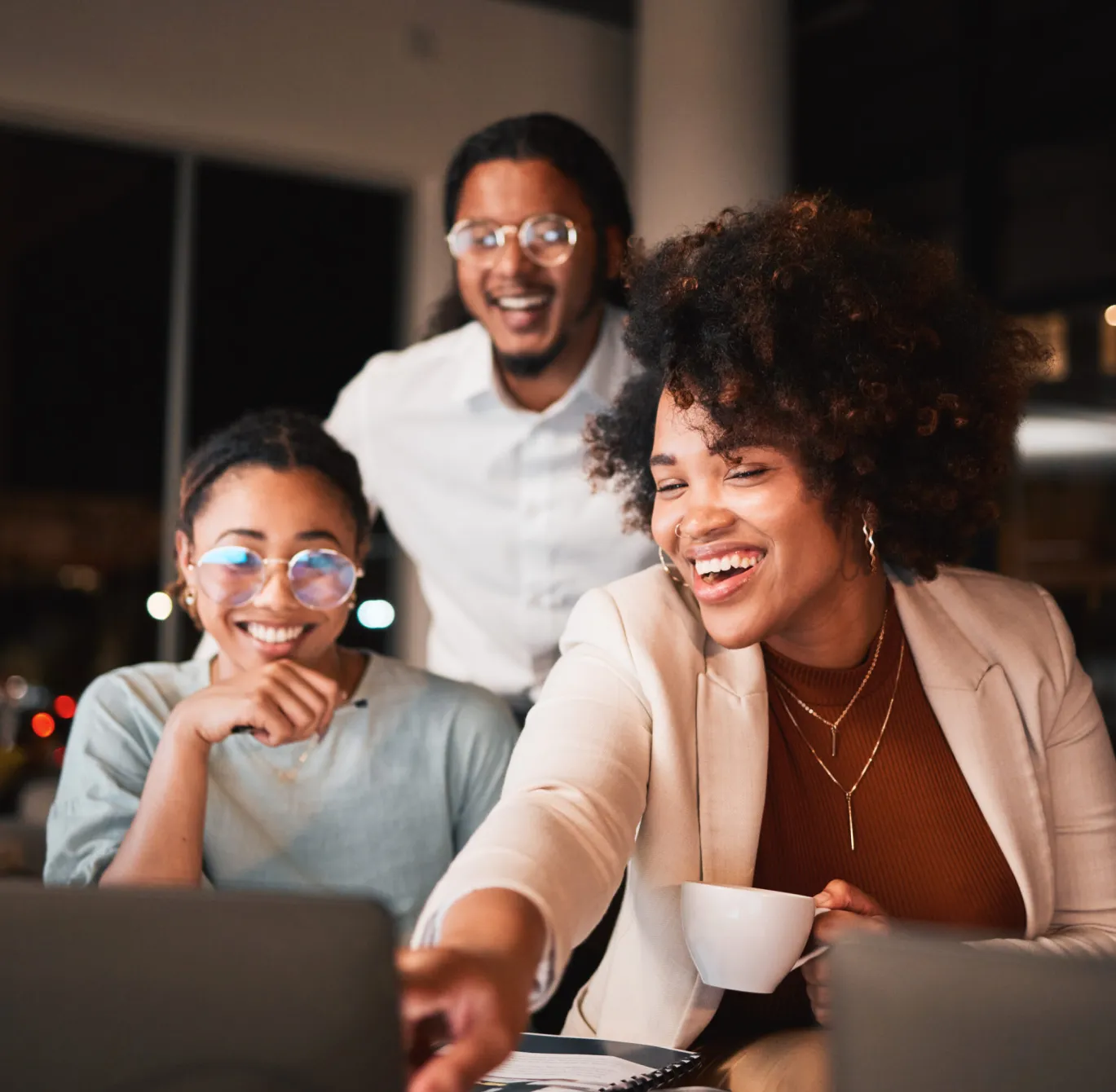 Three people smiling and looking at a laptop together in an office at night.