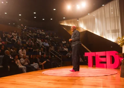 Speaker stands on a TED stage addressing an audience in a theater with warm lighting.