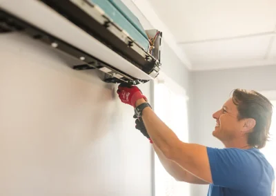 Smiling person in blue shirt repairs wall-mounted air conditioning unit using a screwdriver and red gloves.