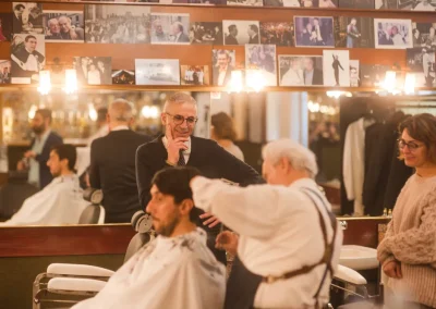 A man gets a haircut in a barbershop, with onlookers and photos displayed on the wall behind them.