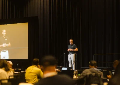 A person speaks on stage at a conference, with attendees seated and watching, and a screen beside them.