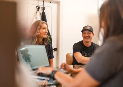 Two people smiling and talking at a table with laptops during a casual indoor meeting.