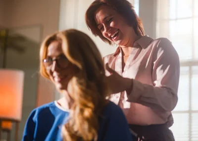 Smiling hairstylist working on a woman’s hair in a sunlit room.