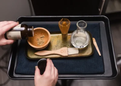 A person prepares a wooden bowl with powder, whisk, brush, and water on a tray for a tea ceremony.