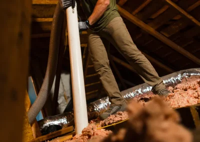 Man in protective gear installs insulation in an attic, holding a large hose with exposed wooden beams above.
