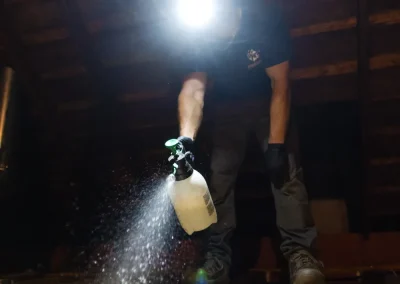 Person wearing a headlamp sprays liquid onto wooden beams in a dimly lit attic space.