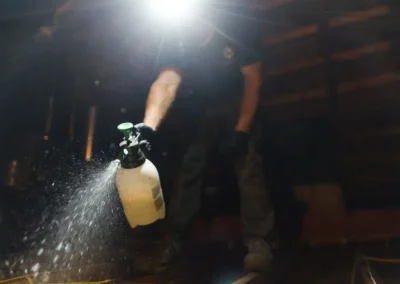 Person spraying liquid from a bottle in a dimly lit attic, wearing gloves and a headlamp.