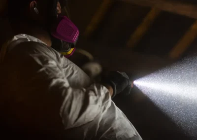 Person in mask and white suit spraying substance into air, highlighting particles in dark setting.