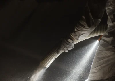 Person in protective suit spraying insulation material from a hose, illuminated by two beams of light.