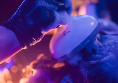 A gloved hand washes hair with a scalp brush under purple lights, creating a vibrant, bubbly scene.